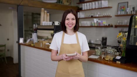 Portrait-of-a-happy-brunette-girl-waitress-in-a-yellow-apron-who-holds-a-notepad-in-her-hands-for-writing-down-orders-in-a-cafe