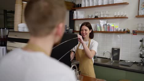 Over-the-shoulder-Happy-brunette-barista-girl-in-a-white-T-shirt-prepares-an-order-for-a-male-waiter-and-communicates-with-him-while-working-in-a-cafe