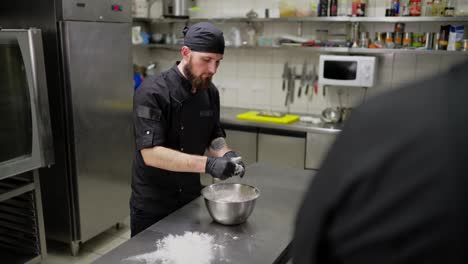 A-confident-male-professional-chef-in-a-black-uniform-transfers-dough-from-a-bowl-to-the-table-and-begins-to-knead-it-before-preparing-pizza-in-a-restaurant-kitchen