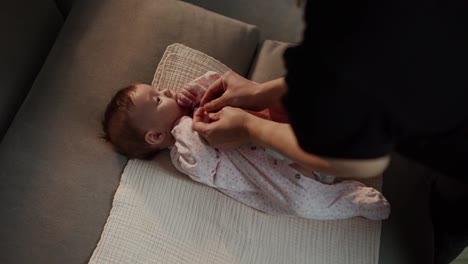 Top-view-of-a-little-girl-baby-lying-on-a-special-mat-on-a-gray-sofa-while-her-mother-fastens-the-buttons-on-the-little-girls-overalls