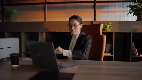 A-concentrated-brunette-girl-in-round-glasses-and-in-a-business-uniform-sits-at-a-wooden-table-and-reviews-sheets-of-papers-with-notes-regarding-the-current-problem-and-its-solution-and-work-plan-in-a-sunny-office