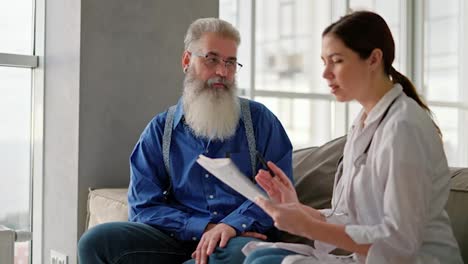 A-brunette-woman-in-a-white-medical-coat-tells-an-elderly-man-with-gray-hair-and-a-lush-beard-in-a-blue-shirt-about-his-need-to-improve-his-health-during-a-home-examination-on-the-sofa-in-a-modern-apartment