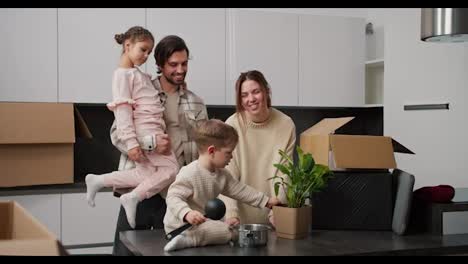 Happy-brunette-girl-in-a-beige-sweater-together-with-her-brunette-husband-in-a-plaid-shirt-who-is-holding-their-little-daughter-in-beige-clothes-talking-while-standing-in-the-kitchen-with-their-little-son-after-moving-to-a-new-apartment