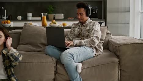 Happy-young-man-with-Black-skin-color-in-a-beige-checkered-shirt-plays-computer-games-on-his-laptop-while-his-young-adult-girlfriend-with-a-bob-hairstyle-does-yoga-while-sitting-on-the-floor-in-a-room-of-a-modern-apartment