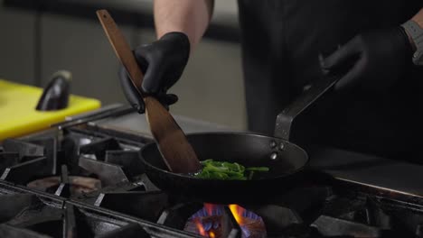 Close-up-a-professional-chef-in-a-black-uniform-fries-green-vegetables-and-adds-white-pieces-of-seafood-to-a-hot-frying-pan-on-a-gas-burner-while-preparing-a-dish-in-a-restaurant-kitchen