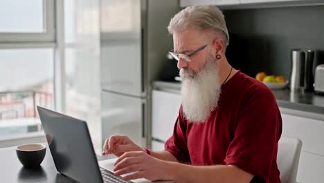 Un-Hombre-Mayor-Con-Gafas,-Cabello-Gris-Y-Barba-Exuberante-Con-Una-Camisa-Roja-Trabaja-En-Su-Computadora-Portátil-Gris-Mientras-Está-Sentado-En-La-Cocina-De-Un-Apartamento-Moderno.