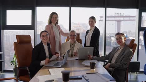 Portrait-of-a-group-of-confident-girls-in-business-suits-wearing-glasses-at-a-table-during-a-meeting-in-a-modern-office-with-large-windows