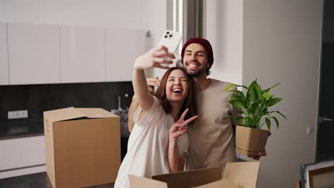 Happy-brunette-girl-in-a-white-T-shirt-takes-a-selfie-using-a-white-smartphone-with-her-boyfriend-with-stubble-in-a-beige-T-shirt-and-house-plants-during-her-move-among-a-large-number-of-boxes-in-a-modern-apartment