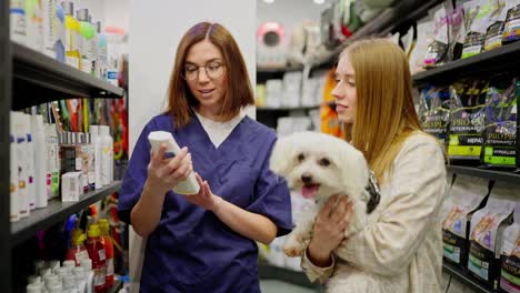 A-confident-brunette-girl-in-a-blue-uniform-an-assistant-in-a-pet-store-talks-about-products-for-a-client-and-her-white-dog