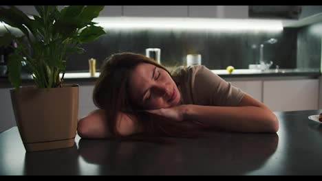 A-brunette-girl-in-a-beige-T-shirt-rests-and-lies-leaning-on-a-black-table-near-a-houseplant-in-a-modern-apartment-in-the-kitchen-with-luminous-ribbons.-Calm-happy-girl-leaning-on-table-near-indoor-flower-at-home-in-the-evening