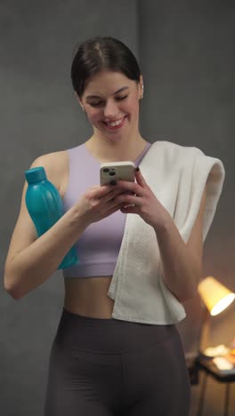 Vertical-video-Confident-and-happy-sporty-brunette-girl-in-a-purple-top-writes-on-social-networks-using-a-white-smartphone-holds-a-white-towel-and-a-blue-sports-bottle-in-her-hands-after-playing-sports-in-a-modern-apartment