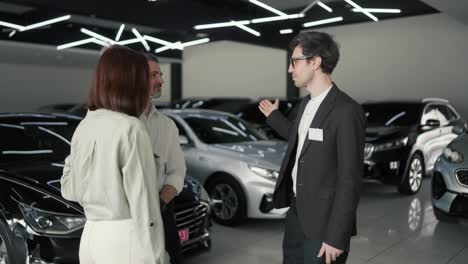 A-confident-male-assistant-in-a-car-dealership-tells-the-client-his-family-his-wife-and-little-son-about-what-cars-are-in-the-showroom-and-about-the-cars.-Confident-young-male-brunette-assistant-in-a-business-suit-talks-about-family-about-cars-in-a-car-showroom