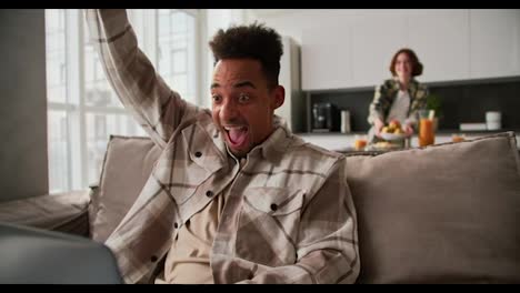 A-happy-and-very-cheerful-young-man-with-Black-skin-and-brunette-is-very-happy-about-his-success-at-work-sitting-at-a-laptop-on-the-sofa-his-girlfriend-runs-up-to-him-and-hugs-him-from-behind-rejoicing-for-her-boyfriend-in-a-modern-apartment