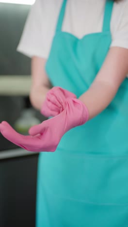 Vertical-video-of-a-girl-in-a-blue-apron-a-professional-cleaner-puts-on-pink-rubber-gloves-before-starting-cleaning-during-a-In-a-modern-apartment-there-is-a-cleaner-on-call