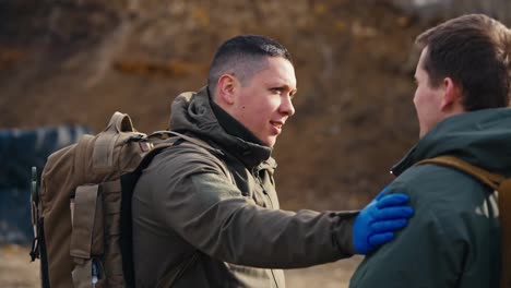 Primer-Plano-Por-Encima-Del-Hombro-De-Un-Hombre-Seguro-De-Sí-Mismo-Con-Un-Corte-De-Pelo-Corto,-Con-Un-Uniforme-Militar-Verde-Oscuro-Y-Guantes-Médicos-Azules,-Le-Da-Una-Palmadita-En-El-Hombro-A-Su-Amigo-Militar-Durante-Su-Comunicación-En-Un-Campo-De-Entrenamiento-Militar-Con-Grandes-Neumáticos-Negros-En-La-Estepa.