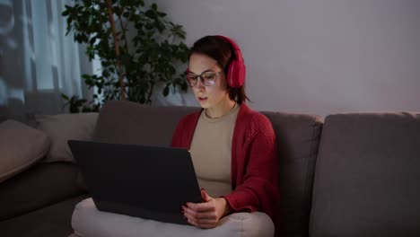 Side-view-of-a-confident-brunette-girl-in-wireless-headphones-and-glasses-sitting-on-a-gray-sofa-and-studying-pronunciation-in-foreign-languages-using-online-lessons-on-gray-laptops-in-a-modern-apartment