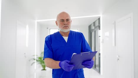 Ein-Männlicher-Arzt-Mit-Brille-Und-Grauem-Bart-In-Blauer-Uniform-Geht-Eilig-Den-Flur-Einer-Hellen-Klinik-Entlang-Und-Hält-Ein-Tablet-In-Den-Händen