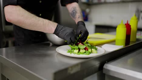 Close-up-a-confident-male-cook-in-black-protective-gloves-puts-a-salad-on-a-plate-and-presents-his-dish-Before-serving-in-a-restaurant-in-the-kitchen