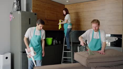 Side-view-of-a-confident-group-of-two-cleaning-guys-and-a-brunette-girl-in-a-white-T-shirt-and-blue-apron-cleaning-in-a-modern-studio-apartment-and-kitchen-on-call