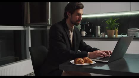 A-confident-brunette-man-in-a-black-jacket-and-beige-shorts-works-at-a-gray-laptop-and-communicates-via-video-in-a-modern-kitchen-in-the-evening.-Confident-brunette-man-with-stubble-communicates-via-video-call-while-sitting-in-only-beige-shorts-in-the-kitchen-in-the-evening