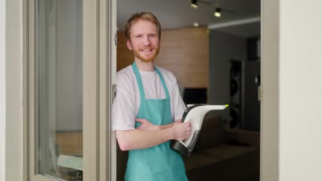 Portrait-of-a-confident-male-blond-cleaner-with-a-beard-in-a-white-T-shirt-and-a-blue-apron-who-holds-a-vacuum-cleaner-in-his-hands-for-cleaning-windows-in-a-modern-apartment