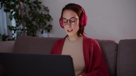 A-happy-and-focused-brunette-girl-in-a-red-sweater-with-glasses-and-wireless-headphones-studies-foreign-languages-using-online-lessons-on-a-gray-laptop-sitting-on-the-sofa-in-a-modern-apartment-in-the-evening