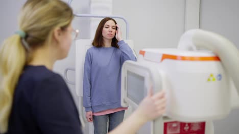 A-confident-brunette-girl-in-a-blue-jacket-stands-near-a-special-fluorography-installation-and-does-fluorography-under-the-supervision-of-a-girl-doctor-nurse-in-a-modern-clinic-in-a-bright-office