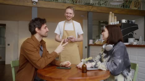 Happy-brunette-guy-in-a-brown-jacket-and-a-girl-in-wireless-headphones-make-an-order-with-a-waiter-in-a-cafe