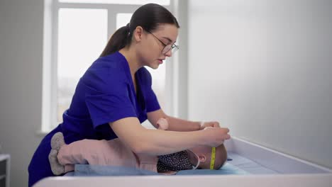 Confident-brunette-girl-with-glasses-in-a-blue-doctors-uniform-measures-the-size-of-a-little-baby-girl-during-prophylaxis-with-pediatricians-in-a-modern-clinic.-Girl-doctor-measures-sizes-and-checks-the-correct-development-of-little-girl-babies-in-a-modern-clinic