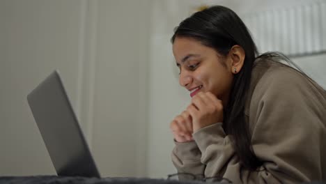 Close-up-of-a-happy-brunette-girl-in-a-brown-sweater-typing-on-her-gray-laptop-while-lying-on-the-bed-in-a-modern-apartment-in-the-bedroom-during-the-day