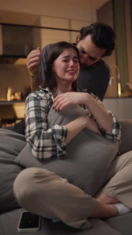 Un-Joven-Moreno-Con-Una-Camiseta-Gris-Calma-A-Su-Novia-Morena-Que-Está-Viendo-Una-Película-Muy-Triste-Y-Llorando-Mientras-Está-Sentada-En-Un-Sofá-Gris-Y-Abrazando-Una-Almohada-En-Un-Apartamento-Moderno-Por-La-Noche.