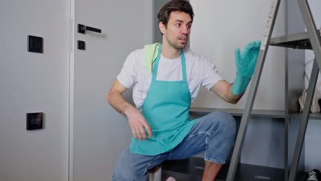 portrait-of-a-confident-happy-male-cleaner-in-a-blue-apron-throwing-a-rag-over-his-shoulder-and-posing-while-sitting-on-the-floor-among-cleaning-tools-and-a-stepladder-in-a-modern-apartment