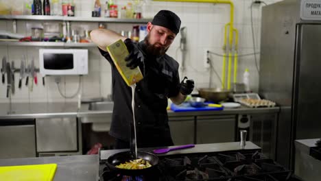 A-professional-male-chef-in-a-black-uniform-pours-cream-over-vegetables-in-a-frying-pan-while-preparing-a-dish-in-the-kitchen-in-a-restaurant