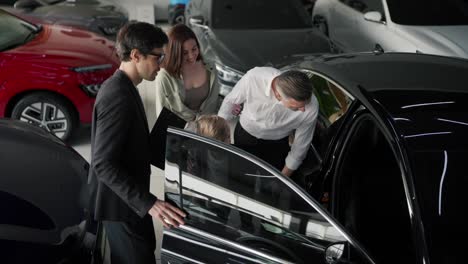 A-family-on-an-excursion-to-a-car-dealership-under-the-supervision-of-a-brunette-man.-An-assistant-in-a-business-suit-a-middle-aged-man-in-a-shirt-together-with-his-son-inspect-the-dealership-and-the-car-from-the-inside