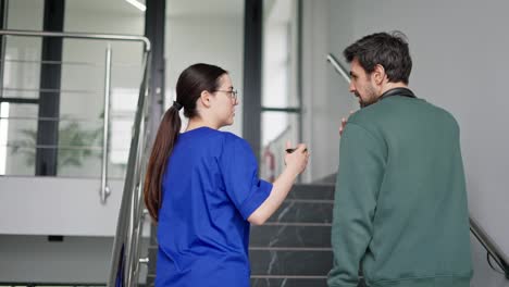 A-confident-brunette-girl-in-round-glasses-and-a-blue-uniform-communicates-with-a-brunette-guy-in-a-green-jacket-about-his-symptoms-while-walking-up-the-stairs-in-a-modern-clinic