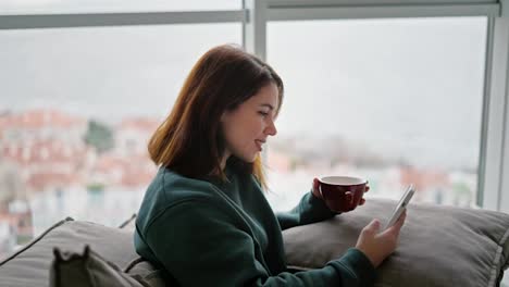 Happy-brunette-girl-on-a-green-sweater-sits-on-a-modern-sofa-drinks-tea-from-a-brown-mug-and-scrolls-through-her-social-media-feed-in-a-White-phone.-Sitting-in-a-modern-apartment-with-large-windows-overlooking-the-sea