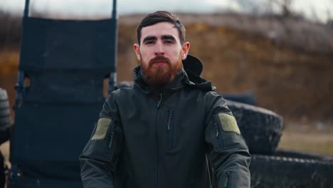 Portrait-of-a-confident-brunette-man-with-a-beard-in-a-dark-green-army-uniform-near-barricades-made-of-rubber-tires-of-a-car-outside-the-city