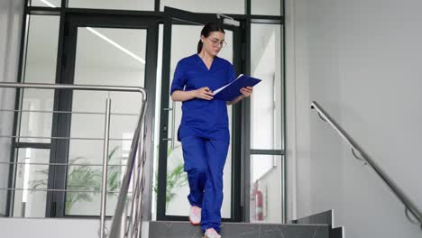 Una-Chica-Morena-Segura-De-Sí-Misma-Con-Gafas-Redondas-Y-Un-Uniforme-Azul,-Un-Médico-Con-Una-Tableta-En-Sus-Manos-Baja-Las-Escaleras-En-Una-Clínica-Moderna.