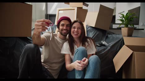 Portrait-of-a-Happy-brunette-man-with-stubble-in-a-red-hat-and-a-beige-T-shirt-who-holds-in-his-hands-the-keys-to-his-new-apartment-sitting-on-the-floor-with-his-girlfriend-near-the-sofa-packed-in-a-black-plastic-case-among-a-large-number-of-boxes-in-his-new-apartment-after-moving