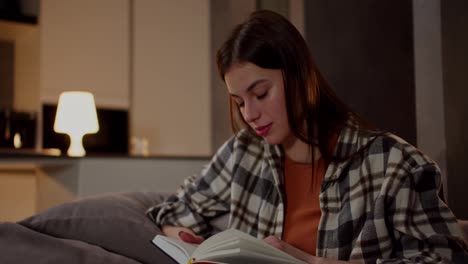 A-happy-brunette-girl-in-a-plaid-shirt-and-an-orange-T-shirt-reads-a-book-and-flips-through-its-pages-while-relaxing-on-a-gray-sofa-in-a-modern-apartment-in-the-evening