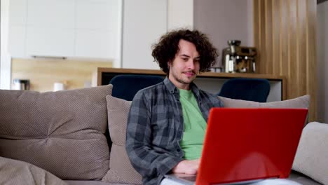 Chico-Moreno-Feliz-Con-Cabello-Rizado-Con-Una-Camisa-A-Cuadros-Gris-Trabajando-En-Una-Computadora-Portátil-Roja-Mientras-Está-Sentado-En-El-Sofá-De-Casa