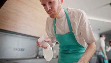 Camera-view-of-a-blond-guy-in-a-white-T-shirt-and-a-blue-apron-as-a-cleaner-washes-the-table-in-the-kitchen-in-a-modern-apartment.-Cleaning-company-in-the-apartment-on-call