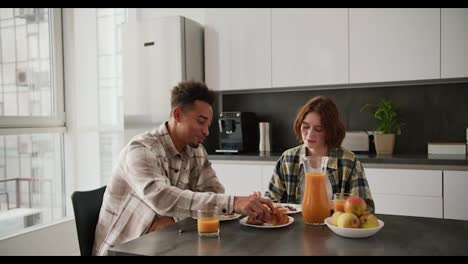 Un-Joven-Moreno-Y-Negro-Feliz-Desayunando-Con-Su-Joven-Novia-Adulta-De-Cabello-Castaño-Con-Un-Peinado-Bob-Y-Una-Camisa-A-Cuadros-Verdes-En-La-Mesa-De-La-Cocina-Por-La-Mañana-En-Un-Apartamento-Moderno