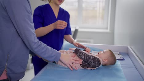 A-confident-and-happy-brunette-mother-and-her-daughter-came-to-the-childrens-pediatrician-for-preventive-care.-Little-girl-baby-lies-in-front-of-girl-pediatrician-doctor-in-modern-clinic