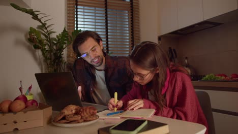 A-confident-and-hardworking-brunette-girl-with-glasses-in-a-red-sweater-studies-foreign-languages-and-writes-notes-in-her-notebook-together-with-a-young-brunette-male-teacher-in-a-modern-apartment-in-the-kitchen