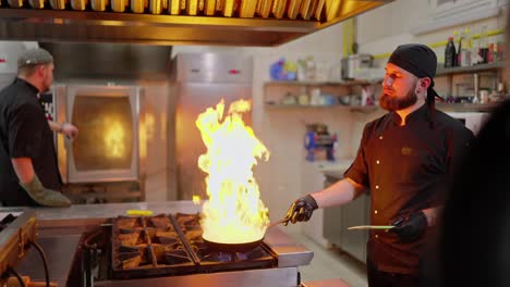 Side-view-of-a-confident-male-chef-in-a-black-uniform-pouring-alcohol-onto-a-hot-frying-pan-with-a-dish-to-use-the-flambéing-technique-and-create-a-controlled-fire-to-give-the-right-taste-to-the-dish-in-the-kitchen