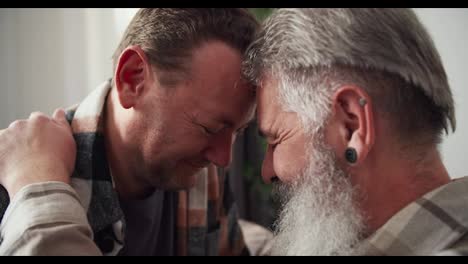 Close-up-an-elderly-man-with-gray-hair-a-lush-beard-and-an-earring-in-his-ear-in-a-checkered-shirt-presses-his-forehead-to-his-brunette-boyfriend-in-a-checkered-shirt-and-holds-him-by-the-shoulder-in-a-modern-apartment-of-an-LGBT-couple