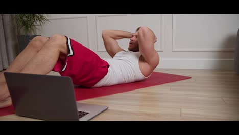 A-confident-blond-man-with-an-athletic-build-and-stubble-in-a-white-T-shirt-and-red-shorts-does-exercises-on-the-mat-to-develop-the-abdominal-muscles-and-watches-a-guide-about-sports-on-his-laptop-in-a-modern-apartment-at-home