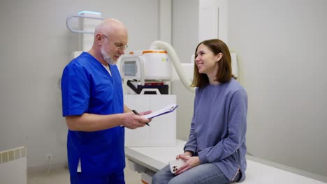 Confident-male-doctor-with-a-gray-beard-in-glasses-and-in-a-blue-uniform-interviews-and-listens-to-the-complaints-of-a-brunette-girl-during-an-appointment-with-a-doctor-in-a-modern-clinic