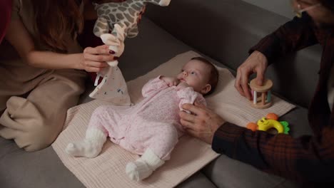 Close-up-of-a-happy-young-brunette-girl-together-with-her-young-husband-playing-with-their-little-daughter-baby-in-a-pink-overalls-with-the-help-of-a-variety-of-toys-for-children-on-a-gray-sofa-in-a-modern-apartment